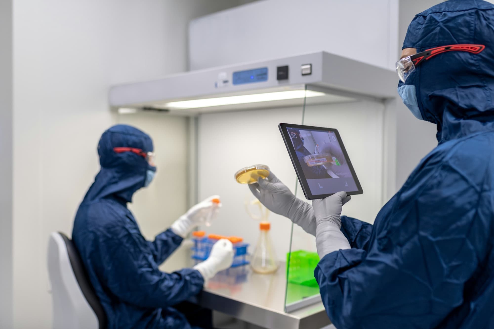 Two scientists in navy suits at a clean-air cabinet; one holds a petri dish, the other a tablet.