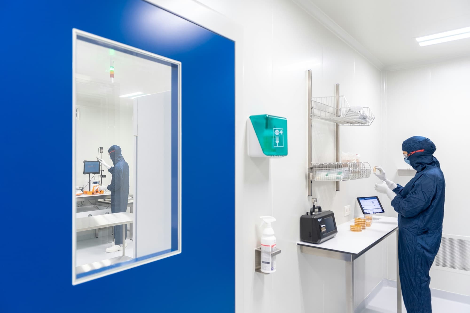 A bright white cleanroom with a royal blue wall and window; a scientist in a navy suit works at a bench.