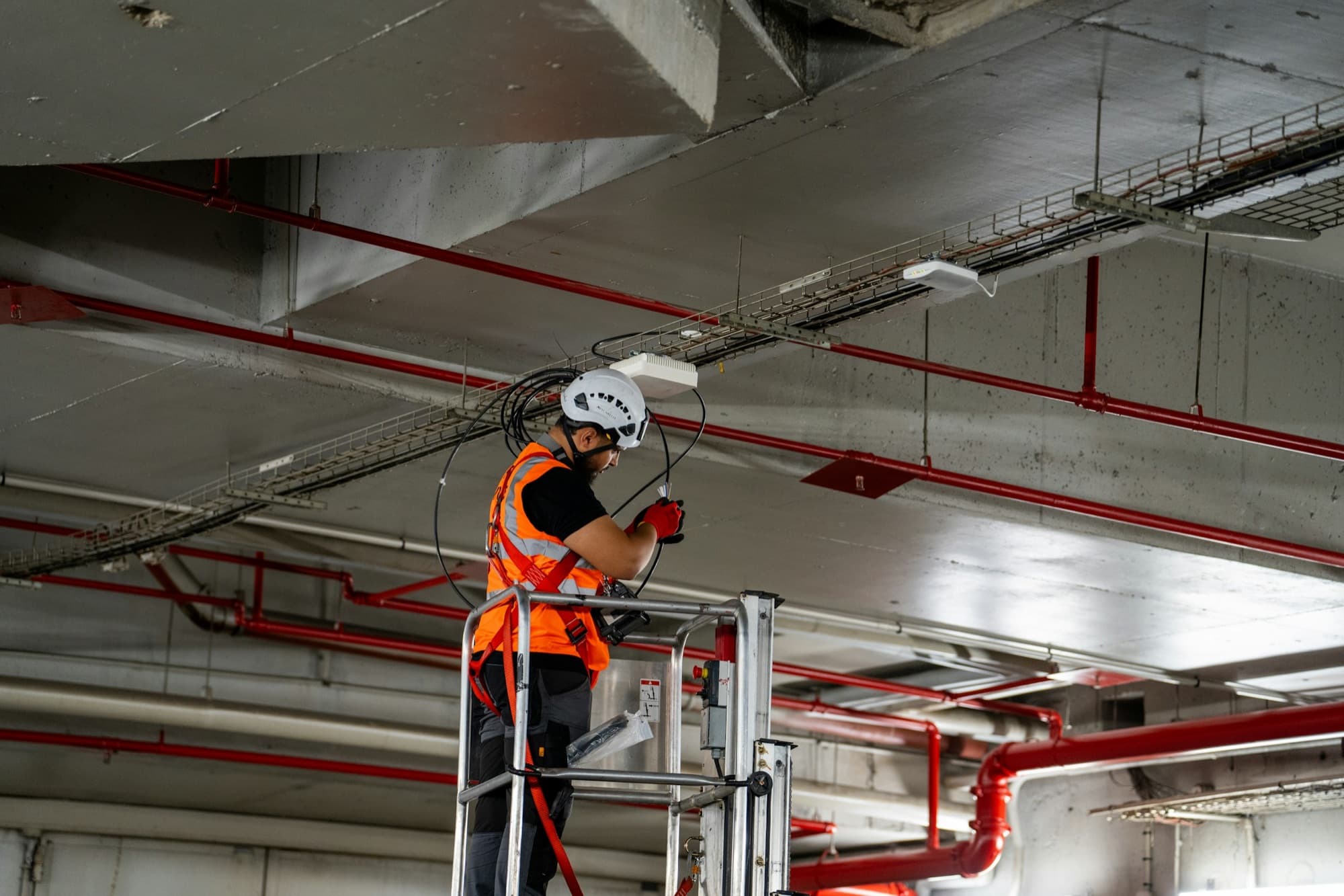 A worker in a hi-vis vest and helmet on a scissor lift installs cabling under a concrete ceiling crowded with pipes and ducts.