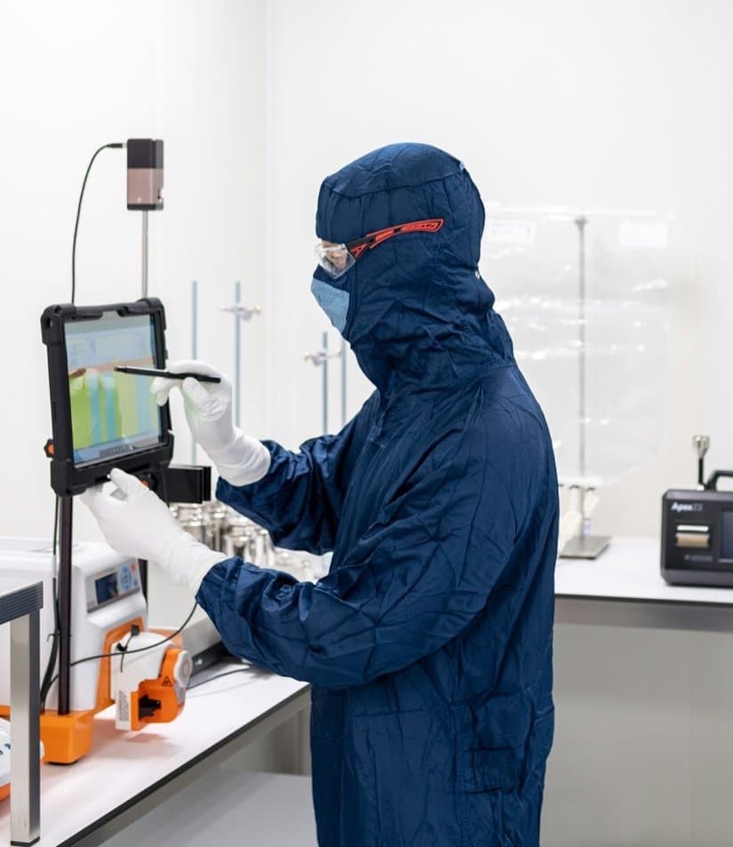 A scientist in a navy cleanroom suit and goggles uses a tablet beside pumps and balances in a bright white laboratory.
