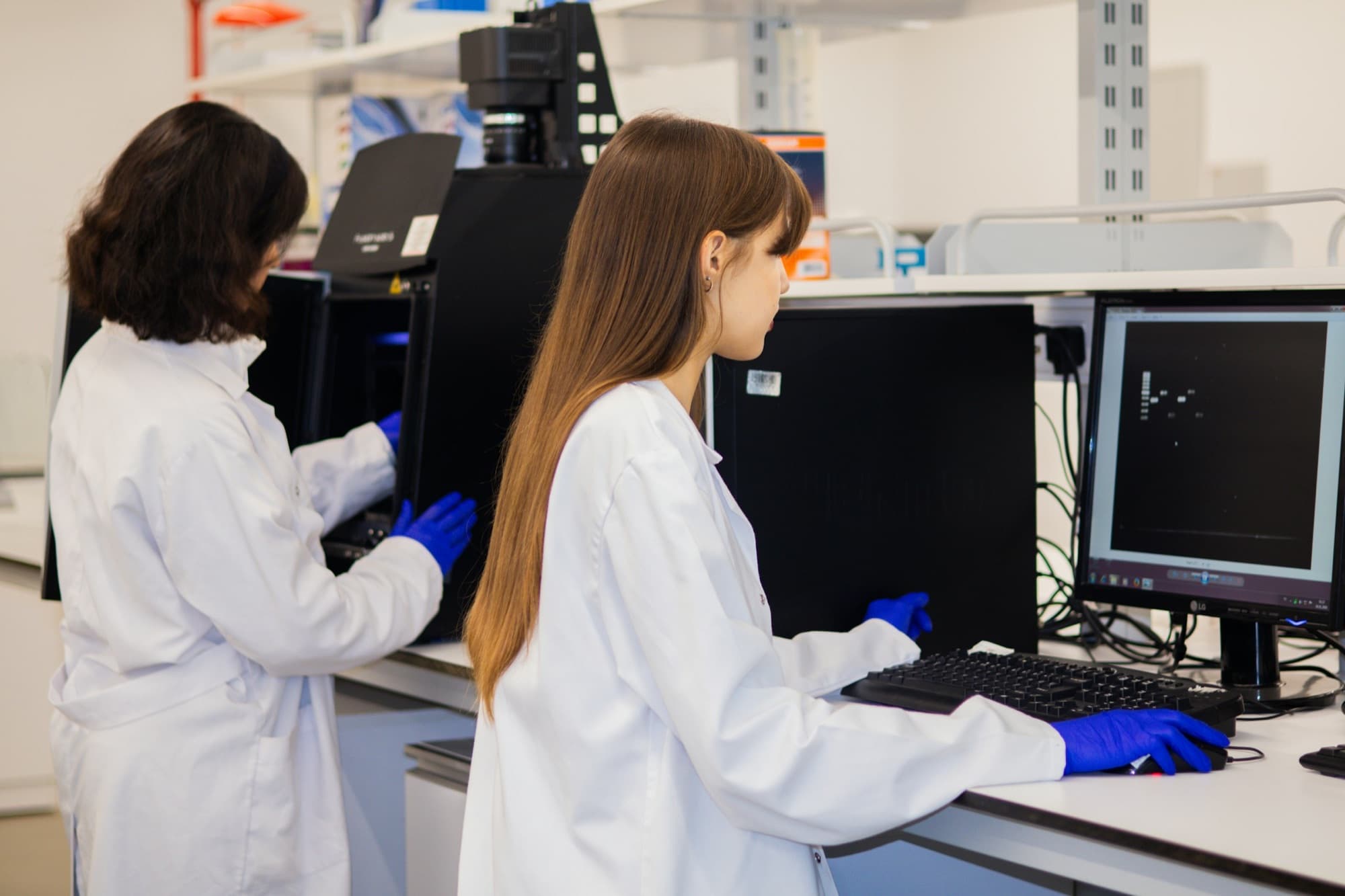 Two scientists in white coats and blue gloves operate a gel imager and computer at a white laboratory bench.