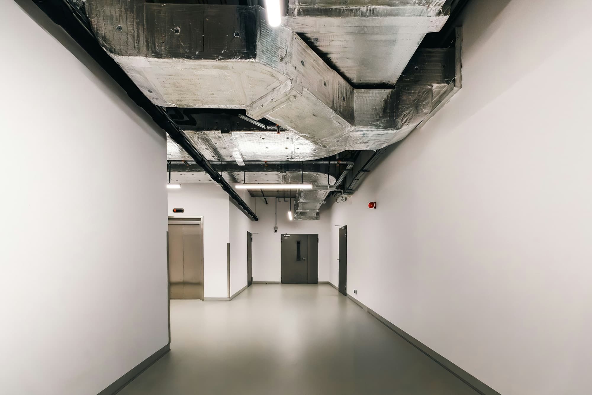 An empty white corridor with exposed, foil-wrapped ductwork and services running overhead.